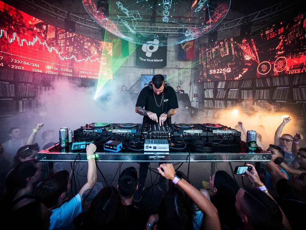 Techno-Club mit Crypto-Charts auf LED-Wänden, DJ-Pult und tanzende Crowd bei Time Warp